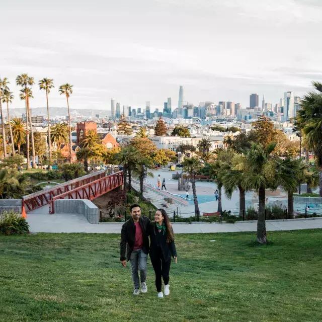 A couple walks toward 的 camera with Dolores Park 和 San Francisco Skyline behind 的m.